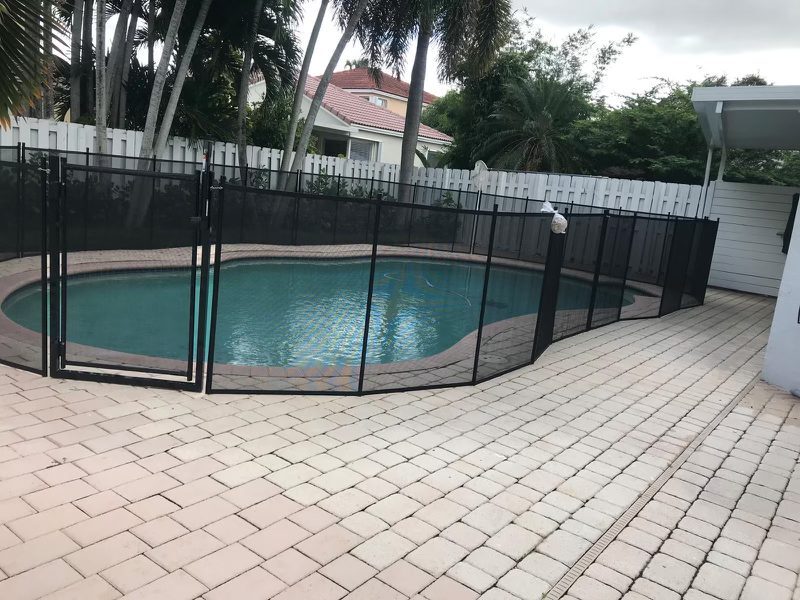 Black mesh fence and gate around a curved pool beside pavers and a long deck drain