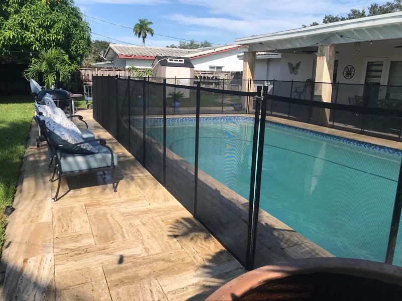 Lowe's pool fence guide illustration: black mesh fence beside a stone walkway and patio seating